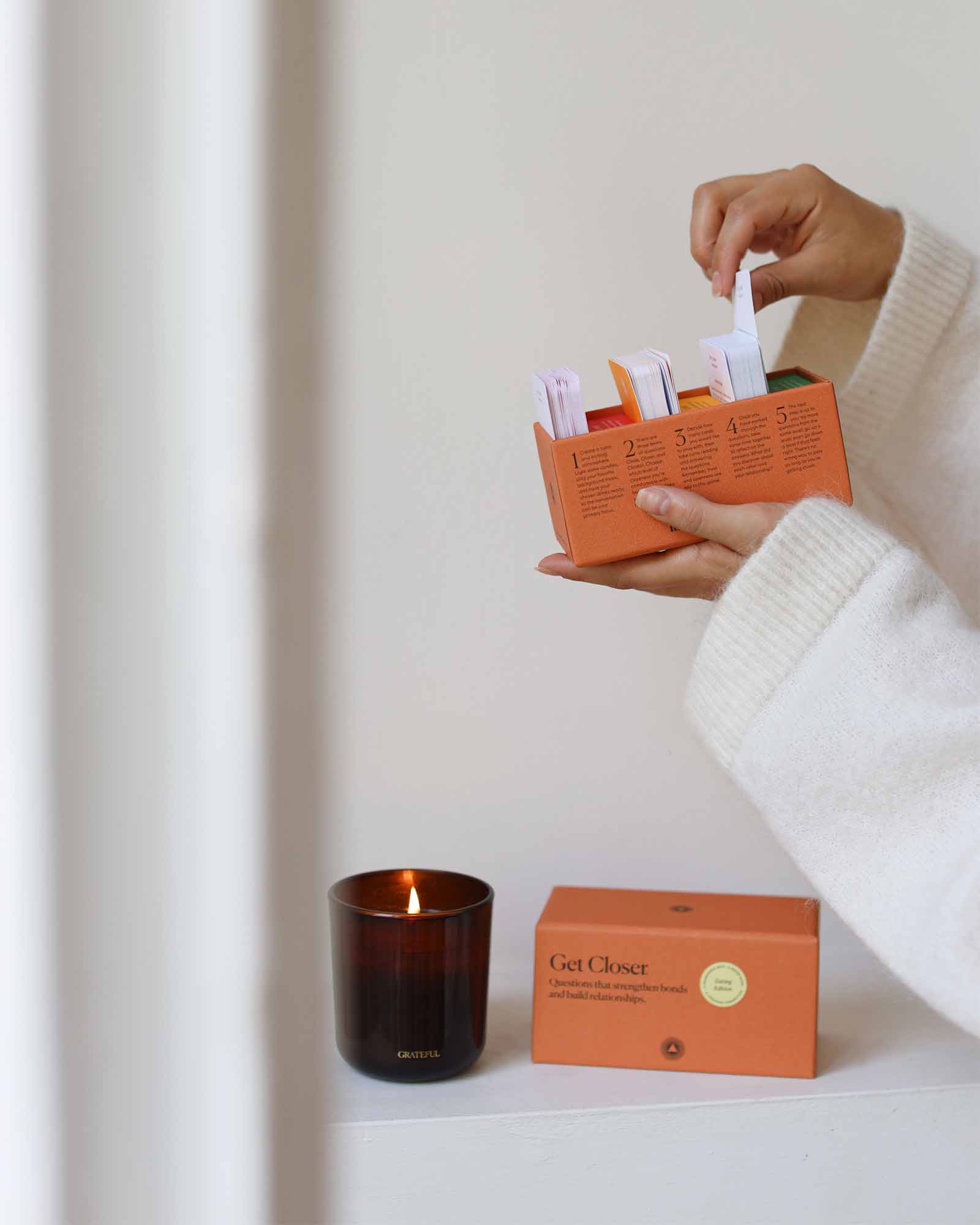 Person holding an orange box with a candle and packaging on a white surface