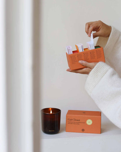 Person holding an orange box with a candle and packaging on a white surface