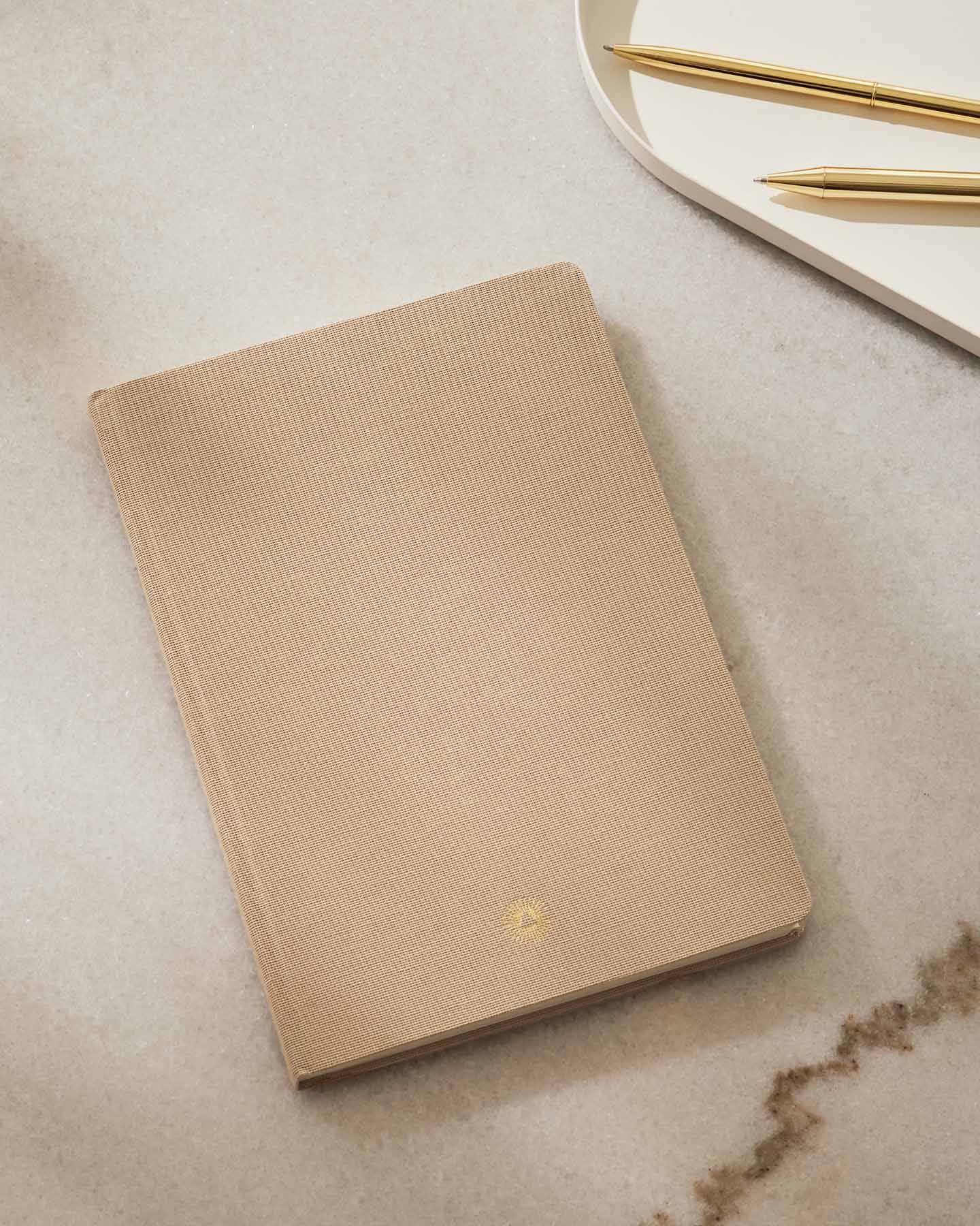 Beige notebook on a marble surface with gold pens in the background