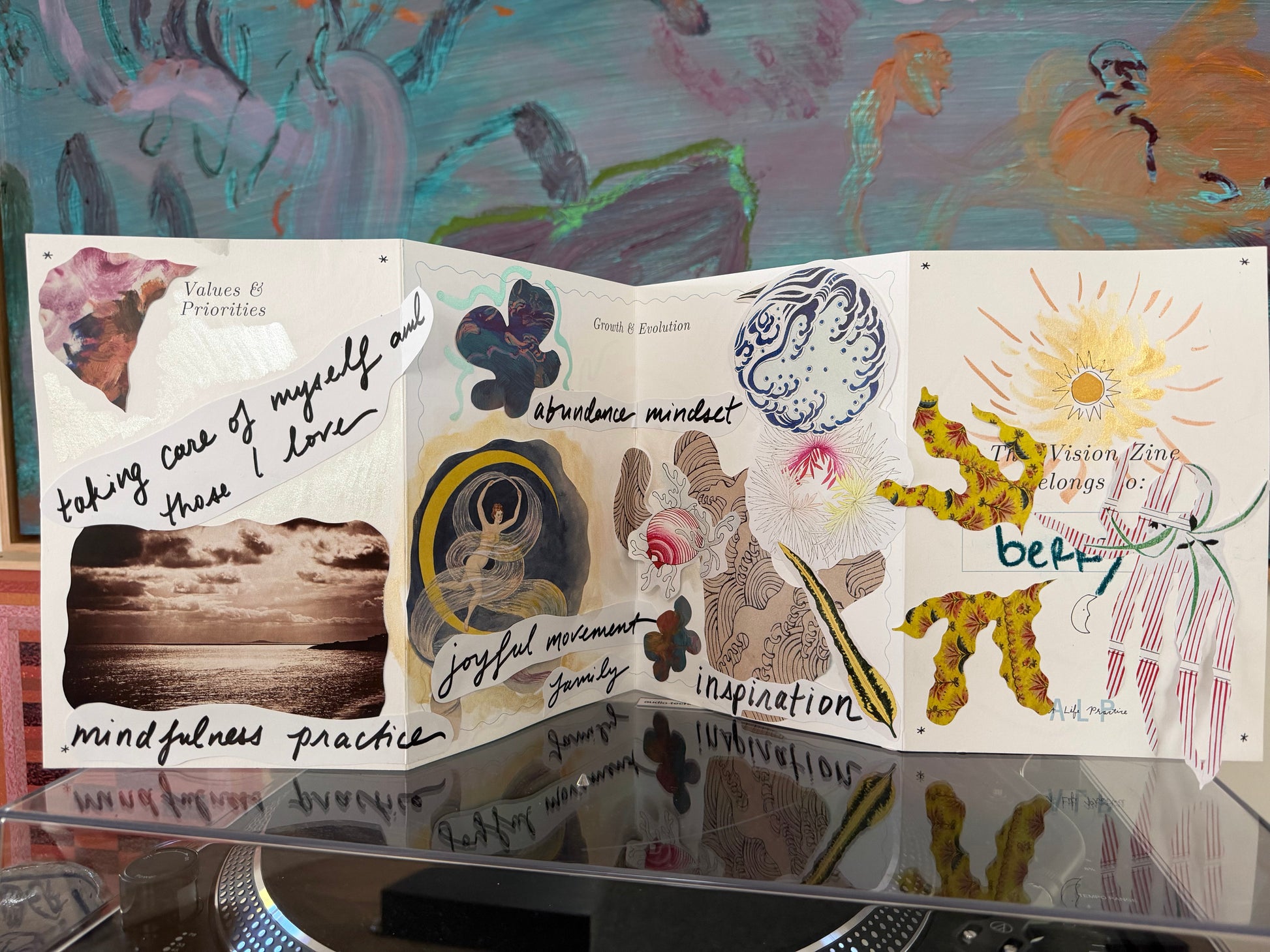 Open book with various illustrations and text on a reflective surface with a colorful abstract painting in the background.