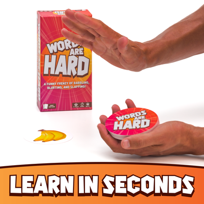 Hand holding a 'Words Are Hard' game piece with packaging in the background on a white background.